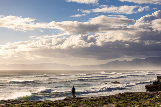 solo female photographer/hiker overlooking cliff with view you massive waves reaching Walker Bay Nature Reserve beach, South Africa at sunset