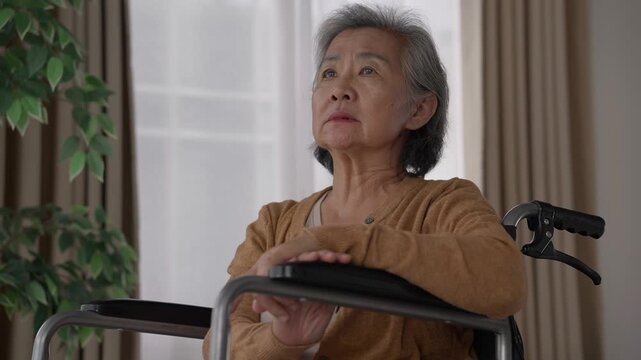 Asian elderly woman sits alone in a wheelchair indoors, looking thoughtful and sad with a heavy sense of solitude and isolation, while facing the window after retirement