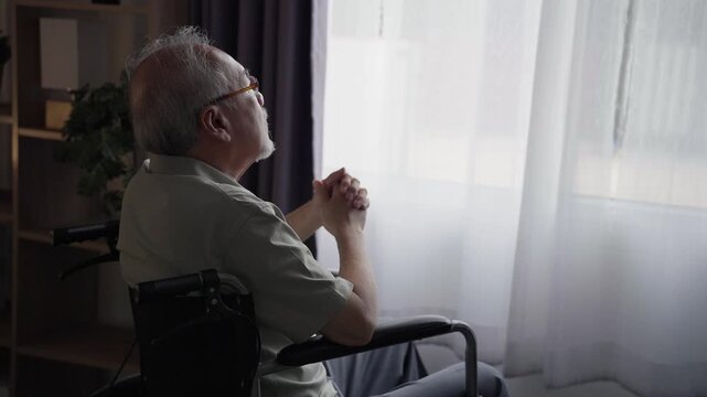 Asian elderly man sits alone in a wheelchair near a window, expressing feelings of loneliness, solitude, and quiet reflection inside a quiet home interior during daytime hours.