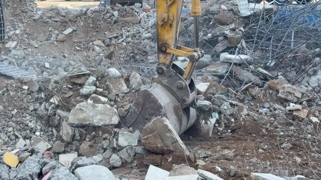 Excavator bucket clears rubble on demolition site. Heavy machine tears through broken concrete and twisted rebar, lifting soil and debris in dusty earthmoving work with relentless industrial power.