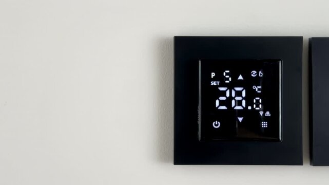 Smart underfloor thermostat turning on to increase domestic heating temperature for synchronization. User sets a higher thermal parameter on the glossy black controller panel. Minimal wall mounted