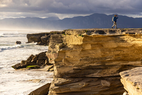 solo female photographer/hiker overlooking cliff with view you massive waves reaching Walker Bay Nature Reserve beach, South Africa at sunset
