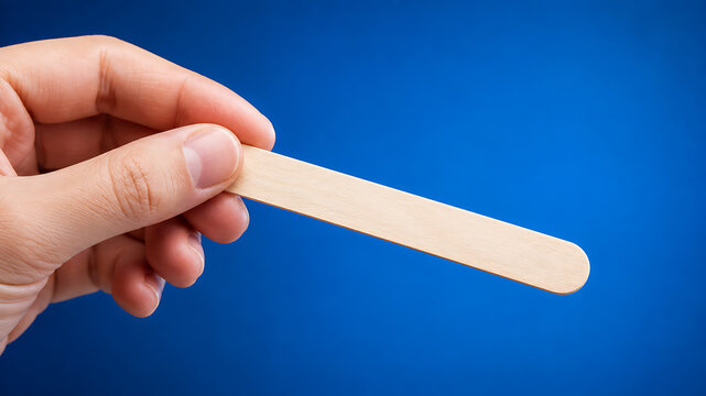 Hand holding wooden tongue depressor against blue background
