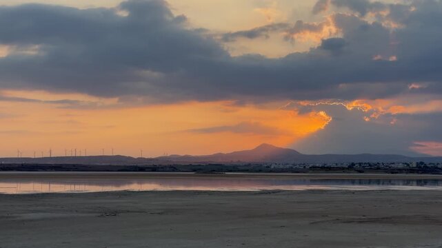 Larnaca Salt lake at sunset glows under layered clouds. Calm water reflects orange light across Cyprus, while distant hills and wind turbines frame quiet landscape in warm evening tones.