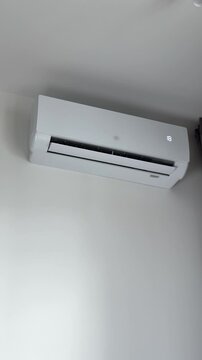 Remote control used by human hand to switch wall conditioner to 17 degrees. Wireless controller regulates severe cooling mode to defeat a hot summer day heat. White indoor split system vent starts