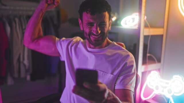 Man holding smartphone, smiling and pumping a fist while seated in a gaming studio with neon controller sign and visible clothing rack; victory joy.