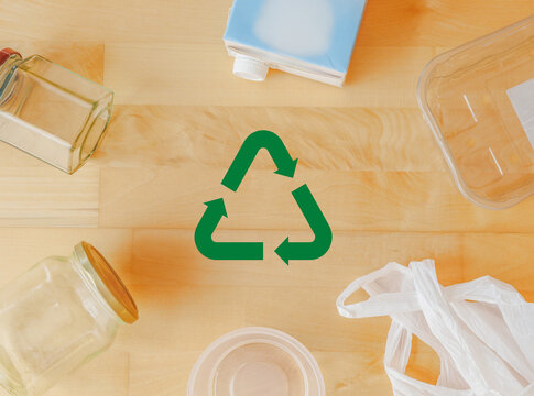 Top view flat lay of household recyclable packaging like glass jars plastic containers and beverage carton surrounding green recycling symbol on wooden background, zero waste and recycling concept