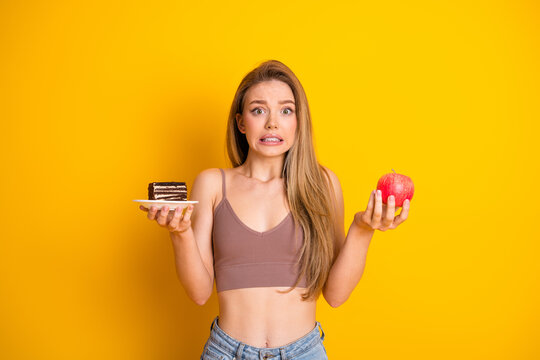 Young woman holding a cake slice and an apple, feeling a dilemma in choosing food options with a yellow background