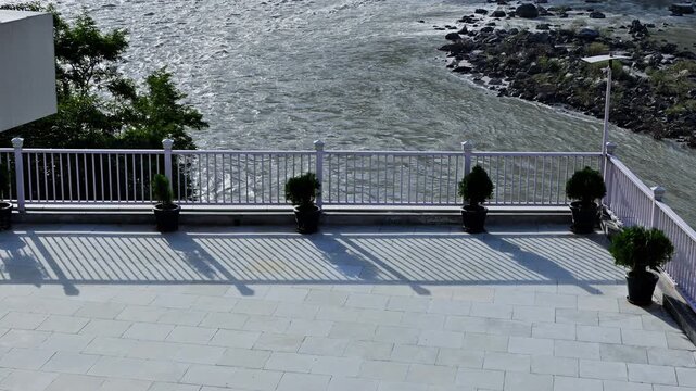 Striped shadows fall across a tiled terrace with potted shrubs and white railings beside the Ganga River in Rishikesh.