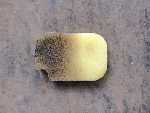 A dirty used yellow cleaning sponge lying on a grey surface.