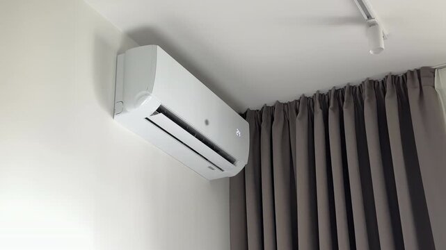 Air conditioning unit functioning silently near deep pleated fabric curtains in a room. White split system horizontal air vent louver moves dynamically for home climate control. Minimalist wall