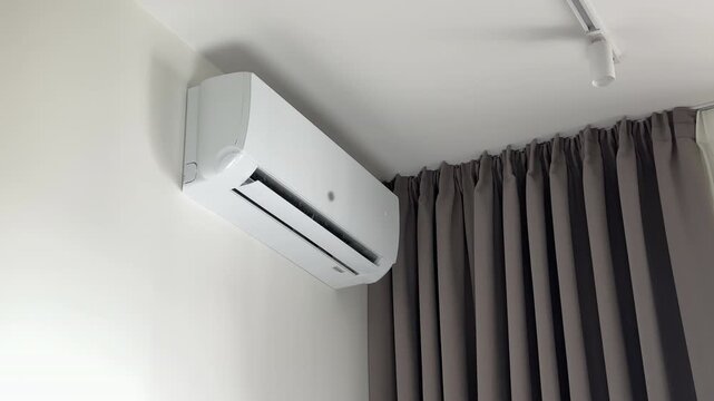 Remote control used by human hand to turn off a wall conditioner. Wireless controller stops the residential climate control device to support power saving routine. White split system appliance shuts