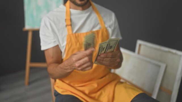 Man painter counting and fanning dollars with hands in studio with easel and canvases visible; satisfaction success.