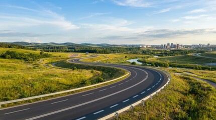 Fototapeta premium A winding highway curves through a lush green landscape towards a distant city skyline. The road is asphalt and bordered by a white safety barrier.