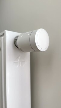 Hand turning on home heating radiator using a white thermostatic controller knob. User sets optimal indoor temperature parameters by adjusting a modern mechanical valve on a white battery. Smart home