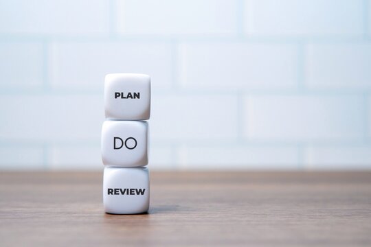Stacked white dice featuring the words PLAN, DO, and REVIEW on a wooden desk. Business management cycles, strategic execution, workflow concept.