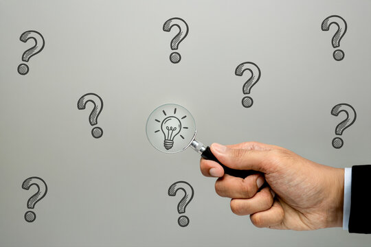 Businessman hand holding magnifying glass revealing lightbulb among drawn question marks on grey background. Search for solution, research, insight, discovery, problem-solving concept.