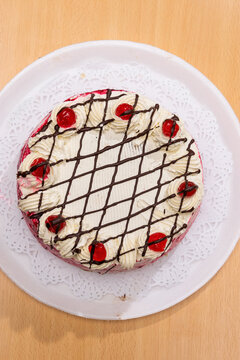 Vista cenital de torta con cobertura blanca, red de chocolate, rosetones de crema y cerezas rojas, sobre fondo de madera.