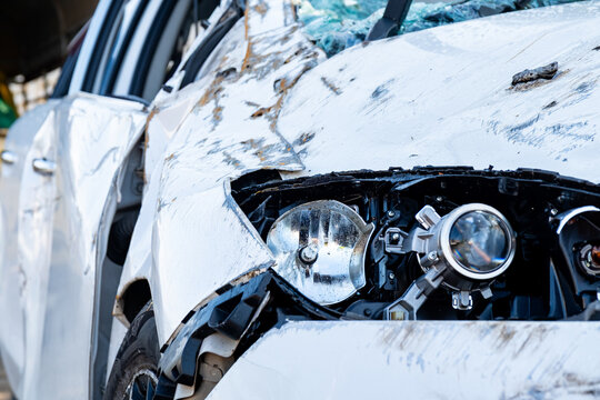 Car accident scene detail with a severely smashed white vehicle headlight. Crushed hood, shattered windshield, and broken car body after a major collision. Focus on impact and wreckage