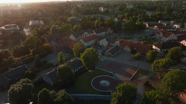 Saldus round plaza and old houses from drone gliding into sunset, Latvia. 4K aerial footage gliding across renovated circular square with green lawn, historic houses, red tiled roofs, trees.