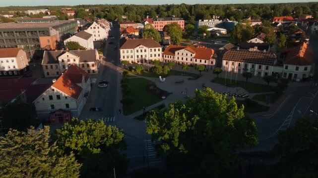 Saldus town square with flower beds from descending drone, Latvia. 4K aerial footage gliding over treetops down to paved plaza with flower beds, lawns, white sculpture, historic buildings.