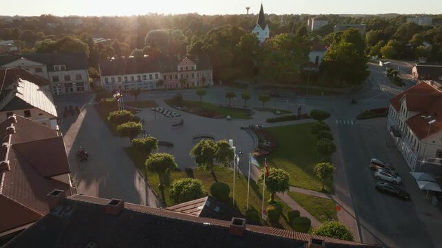 Saldus town square with flagpoles and church spire from rising drone, Latvia. 4K aerial footage climbing above red roofs to reveal paved plaza, lawns, flower beds, trimmed trees.