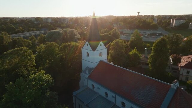 Historic white church with black steeple from orbiting drone at sunset. 4K aerial footage circling old lutheran church with black spire, red tiled roof.