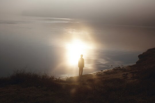 Silhouette standing on shoreline with glowing auric light reflection over calm water for spiritual background and meditation poster