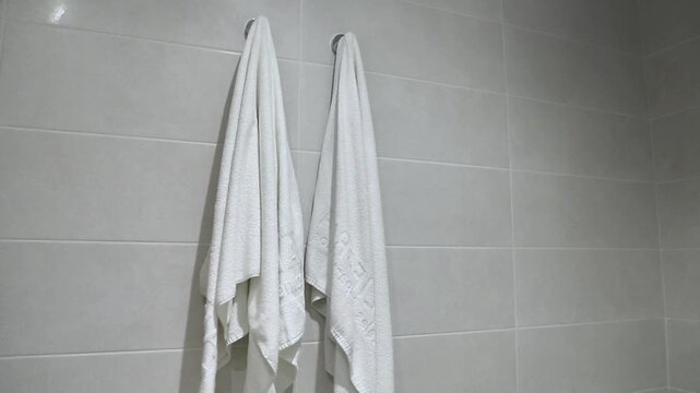 Two white towels hanging on hooks in a bathroom, showing gradual movement and slight repositioning against a light gray tiled wall with a clean and modern aesthetic