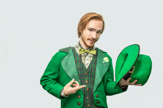 Young readhead man in green suit hold hat in hand and point on it. Guy look on camera seriously. Isolated on grey background.