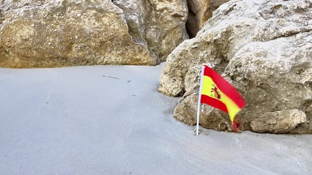Spanish flag positioned among rocks on sandy beach, showcasing vibrant colors of red and yellow, with natural textures of stone and sand creating a serene coastal atmosphere