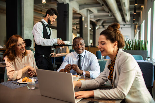 Business colleagues reviewing laptop while waiter serves coffee in cafe