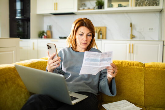 Woman checking bill with smartphone and laptop at home