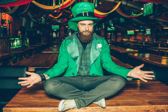 Nice handsome young bearded man in St. Patrick's suit sit with legs crossed on table in pub. His costume green. Guy pose.