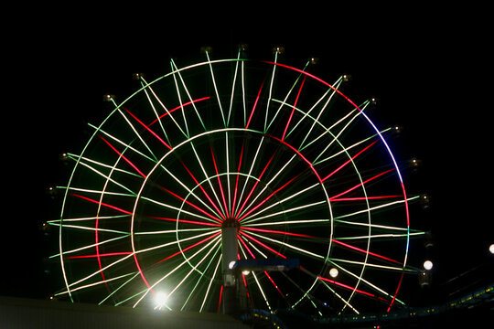 A Ferris wheel illuminated with red and green lights against a black night sky. Bold glowing spokes form a striking geometric wheel that stands out against the darkness.