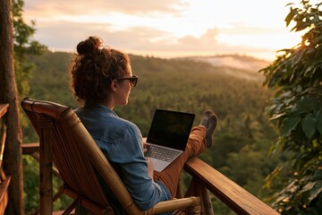 Obraz premium Woman using laptop on bamboo chair overlooking tropical forest at sunset