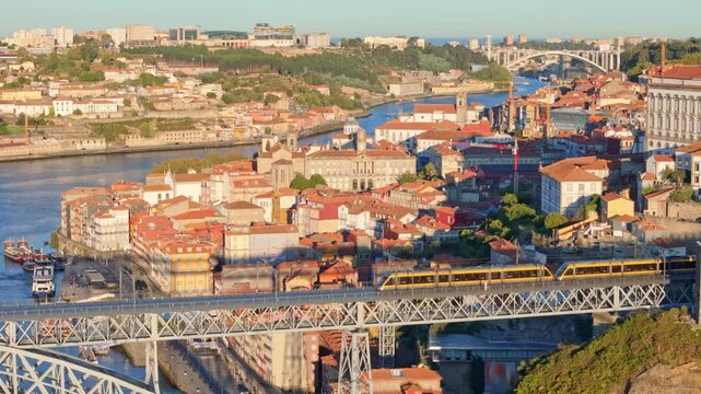 Drone view of Dom Luis I Bridge over Douro River in Porto, Portugal, with lower deck lines, including a tram on the bridge and rabelo boats by the quay at sunrise. Pan shot
