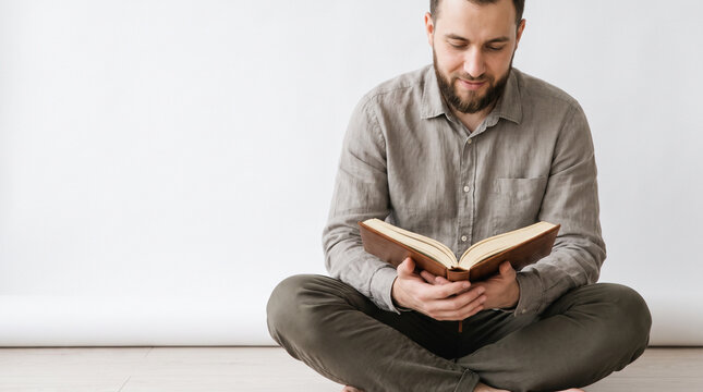 Man reading the Quran while seated cross-legged indoors, with a peaceful downward gaze, neutral clothing and bright white copy space, expressing Ramadan reflection, worship and faith.