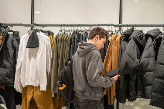 Boy lifts sleeve of warm puffer jacket examining winter collection, customer checking fabric before buying, scene every shop assistant knows. Wide shot. Concept of decision of young customer at rack