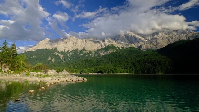Wide static video of the Eibsee shoreline with clear water and the towering Zugspitze mountain range under a cloudy sky in Germany.