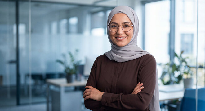 Professional woman wearing a hijab and glasses standing with arms crossed in a modern office environment