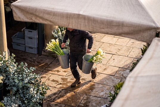 A male florist carries galvanized metal buckets filled with fresh white calla lilies across a stone paved flower market