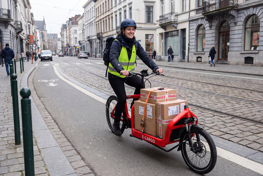 Female Courier Riding Electric Cargo Bike Delivering Packages on City Street in European Town