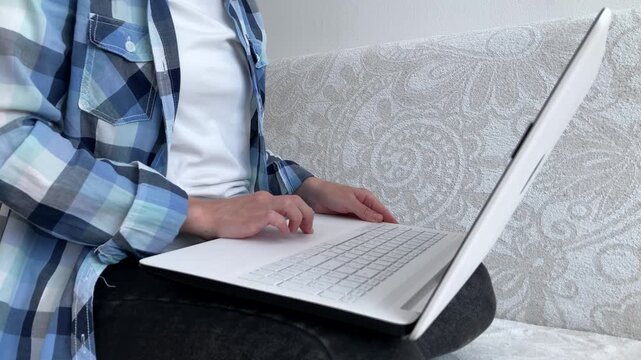 A female freelancer is completing work tasks on a laptop while sitting on a sofa in casual clothes. The topic of remote work, online employment, and the possibility of working from home. Side view