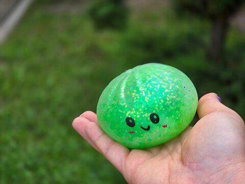 green glitter kawaii squishy toy with cute smiley face and rosy cheeks held in hand, sparkly round ball squished outdoors