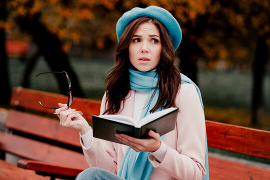 Surprised woman reading book on park bench in autumn wearing blue beret and scarf holding glasses