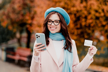 Obraz premium Young woman smiling while shopping online with smartphone and credit card in autumn park