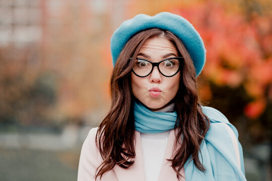 Playful young woman in blue beret and glasses making a surprised kissy face outdoors in autumn fashion portrait