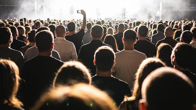 A large crowd of people at a concert, illuminated by stage lights, with one person in the foreground holding up a phone to record.