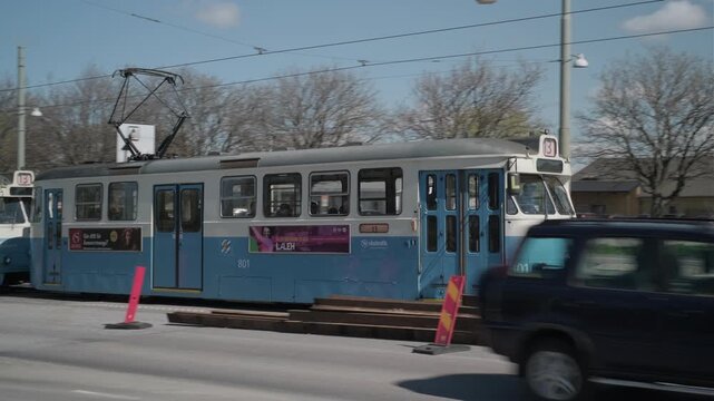 Gothenburg, Sweden - May 2 2023: A vintage blue and white tram crosses a bridge in traffic, with the Red Building (R&ouml;dakon/Kanonhallen) and Gothenburg's skyline visible in the background under a clear